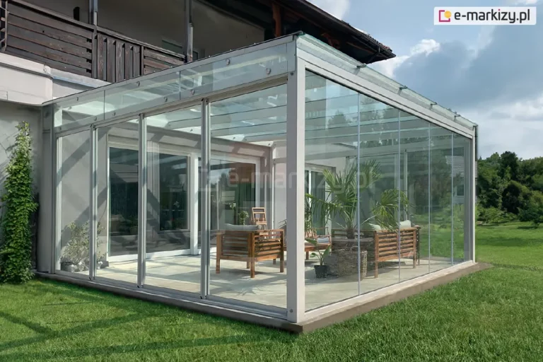 Przyścienna pergola ogrodowa o funkcji ogrodu zimowego, zlokalizowana na terenie zielonym. Konstrukcja opiera się na aluminiowym stelażu w białym kolorze, z dachem wykonanym z hartowanego szkła montowanego punktowo. Ściany obiektu stanowią systemowe przeszklenia ramowe Sotro, które umożliwiają swobodne przesuwanie segmentów szklanych w celu otwarcia przestrzeni. Całość posadowiona jest na betonowym fundamencie wystającym nieznacznie ponad poziom zadbanego trawnika. Wewnątrz przeszklonego pomieszczenia zaaranżowano strefę wypoczynku z drewnianymi meblami ogrodowymi, sofami z szarymi poduchami oraz roślinnością doniczkową, w tym palmą. W tle widoczna jest elewacja piętrowego budynku z drewnianymi balustradami balkonowymi oraz rozległy ogród z drzewami i krzewami. Transparentność zastosowanego systemu szklenia zapewnia niezakłócony widok na otaczający krajobraz przy jednoczesnej ochronie wnętrza przed warunkami atmosferycznymi.