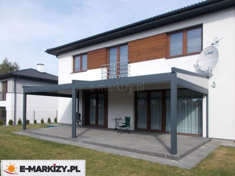 Zdjęcie przedstawia nowoczesną pergolę rzymską Moderno w wersji przyściennej, zamontowaną na dużym, podwyższonym tarasie wyłożonym szarymi płytami. Konstrukcja pergoli składa się z grafitowego, aluminiowego stelaża oraz rozsuwanego, jasnego poszycia dachowego. Zadaszenie jest oparte na trzech solidnych, pionowych słupach o kwadratowym przekroju, które stabilizują konstrukcję od strony ogrodu. System zamontowany jest pod balkonem piętrowego domu o białej elewacji z elementami drewnianej szalówki. Pergola osłania szerokie przeszklenia tarasowe i okna w brązowych ramach, zapewniając cień w strefie wypoczynkowej, gdzie widoczne są pojedyncze meble ogrodowe, takie jak krzesło turystyczne i stolik. Wokół tarasu rozciąga się zadbany trawnik, a w tle widać sąsiednią zabudowę jednorodzinną.