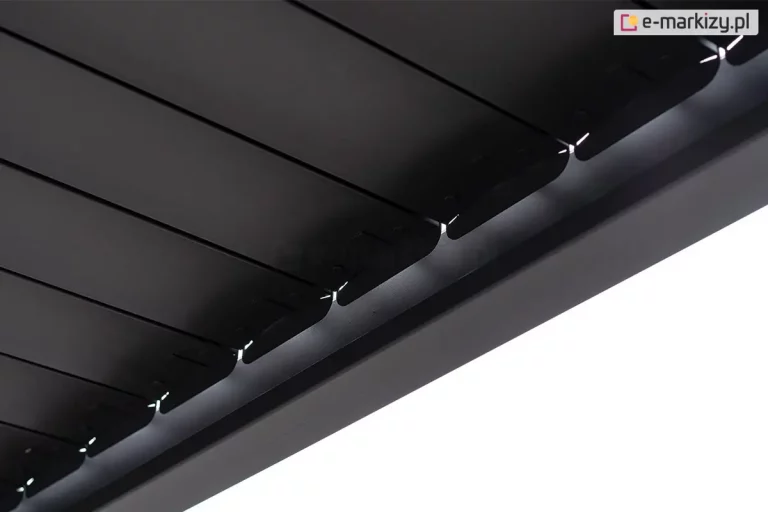 zbliżenie na techniczny detal konstrukcyjny dachu bioklimatycznej pergoli lamelowej Gloria w kolorze antracytowym. Fotografia wykonana z perspektywy dolnej ukazuje rzędowe ułożenie aluminiowych lameli w pozycji niemal całkowicie zamkniętej, co demonstruje szczelność i precyzyjne dopasowanie poszczególnych elementów osłonowych. Widoczne są krawędzie lameli stykające się z wewnętrznym profilem ramy bocznej, która pełni funkcję rynny odprowadzającej wodę opadową. Powierzchnia aluminium posiada matowe wykończenie o drobnej strukturze, a w miejscach styku piór z ramą widoczne są systemowe uszczelnienia oraz punkty montażowe odpowiedzialne za stabilność i rotację dachu. Kontrastujące światło przebijające przez minimalne szczeliny montażowe przy krawędzi ramy podkreśla solidność i masywność profilu nośnego pergoli.