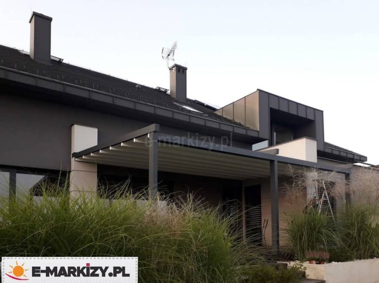 Zdjęcie prezentuje nowoczesną pergolę rzymską Moderno w wersji przyściennej, zainstalowaną na tarasie domu o ciemnej, grafitowej elewacji. Aluminiowa konstrukcja pergoli w kolorze antracytowym współgra z barwą ścian budynku oraz dachu krytego blachą na rąbek stojący. Zadaszenie składa się z białego, rozsuwanego poszycia z tkaniny, która po całkowitym rozłożeniu tworzy płaską powierzchnię z lekkim nachyleniem. Całość wspiera się na dwóch solidnych profilach pionowych zamontowanych do podłoża tarasu. W dolnej części kadru widoczna jest gęsta roślinność w postaci ozdobnych traw, które częściowo zasłaniają strefę wypoczynkową. Dom posiada nowoczesną bryłę z dużymi przeszkleniami oraz pionowymi akcentami elewacyjnymi w kolorze beżowym, co tworzy kontrast z ciemną bazą budynku.