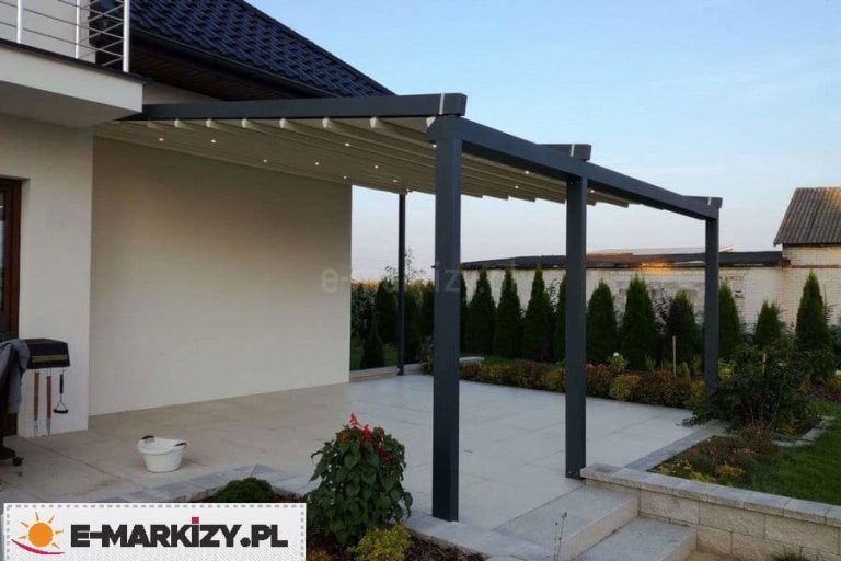 Zdjęcie prezentuje nowoczesną pergolę rzymską Moderno w wersji przyściennej, zainstalowaną na dużym tarasie wyłożonym jasnymi płytami wielkoformatowymi. Konstrukcja pergoli wykonana jest z profili aluminiowych w kolorze ciemnoszarym (antracytowym), a jej dach tworzy rozsuwane poszycie z białej tkaniny technicznej. W poprzecznych profilach podtrzymujących materiał zintegrowano punktowe oświetlenie LED, które jest widoczne pod zadaszeniem. System jest zamontowany jedną krawędzią do jasnej elewacji piętrowego budynku, natomiast przednia część opiera się na trzech masywnych nogach o kwadratowym przekroju. Całość posiada wyraźny spadek połaci dachowej w kierunku ogrodu, co ułatwia odprowadzanie wody. W tle widoczny jest zadbany trawnik, pas zielonych tui oraz fragmenty niskiej roślinności ozdobnej przy schodach prowadzących z tarasu do ogrodu.