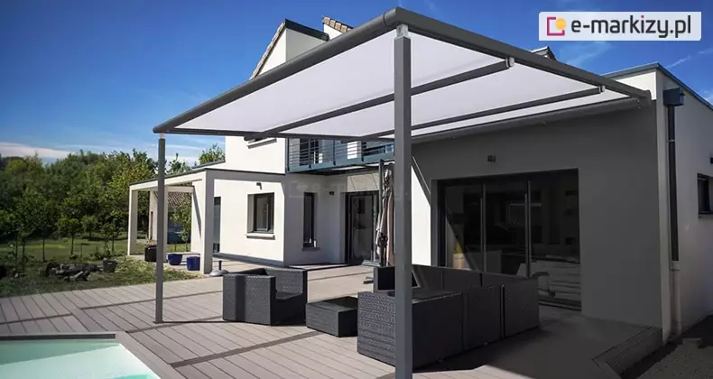 Nowoczesna pergola tarasowa przyścienna V Ziiip Aluprof, wykonana z profili aluminiowych w kolorze antracytowym. Zadaszenie stanowi biała, półtransparentna tkanina techniczna, rozpięta na systemie prowadnic, która osłania strefę wypoczynkową przed słońcem. Konstrukcja opiera się na dwóch kwadratowych słupach nośnych, zamontowanych na tarasie wykonanym z jasnobrązowych desek kompozytowych ułożonych równolegle. Pod pergolą znajduje się zestaw mebli ogrodowych z ciemnego technorattanu, w tym narożna sofa, fotel oraz stolik kawowy. W tle widoczna jest bryła nowoczesnego, piętrowego domu o białej elewacji z ciemnymi obramowaniami okien oraz szklanymi drzwiami przesuwnymi prowadzącymi do wnętrza. Po lewej stronie kadru znajduje się fragment basenu z jasnoniebieską wodą, a w dalszym planie zadbany ogród z trawnikiem i drzewami liściastymi pod czystym, niebieskim niebem.