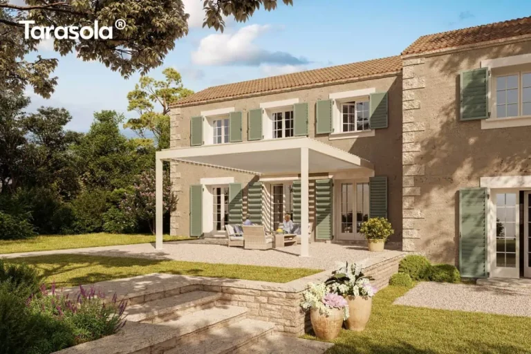 Zdjęcie przedstawia wizualizację luksusowej rezydencji w stylu śródziemnomorskim z zamontowaną nowoczesną konstrukcją Essential tarasola w kolorze kremowym lub jasnobeżowym. Budynek o piaszczystej, kamiennej elewacji posiada liczne okna i drzwi balkonowe ozdobione klasycznymi, drewnianymi okiennicami w kolorze stonowanej zieleni. Pergola o minimalistycznej formie wsparta jest na dwóch smukłych słupach i przylega bezpośrednio do ściany domu, osłaniając przestronny taras wysypany jasnym żwirem. Pod zadaszeniem z białych lameli znajduje się strefa wypoczynkowa z wygodnymi, beżowymi fotelami i sofą oraz niskim stolikiem kawowym. Na tarasie ustawiono również dużą donicę z kwitnącym krzewem. Posesja jest otoczona gęstą roślinnością i drzewami, a na pierwszym planie widoczne są szerokie, kamienne schody prowadzące do ogrodu, przy których stoją duże, gliniane wazy z białymi i różowymi kwiatami. Całość skąpana jest w ciepłym świetle słonecznym, tworząc atmosferę spokoju i elegancji w letnie popołudnie.