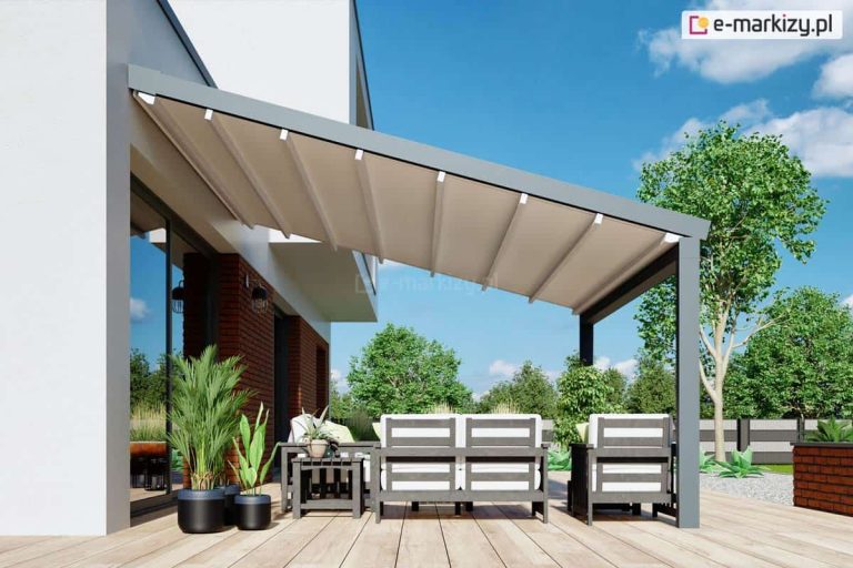 Nowoczesna pergola przyścienna Moderno o aluminiowej konstrukcji w kolorze antracytowym, zamontowana nad tarasem wykonanym z jasnych desek kompozytowych. Zadaszenie stanowi ruchoma tkanina techniczna w kolorze kremowym, która jest w pełni rozwinięta i zamontowana na prowadnicach pod kątem, umożliwiając swobodny odpływ wody. Pod pergolą zaaranżowano strefę wypoczynkową z szarymi meblami ogrodowymi o drewnianej konstrukcji, białymi poduszkami oraz czarnymi donicami z roślinnością. Konstrukcja przylega do białej elewacji budynku z elementami czerwonej cegły i dużymi przeszkleniami, a w tle widoczne są zielone drzewa oraz ogrodzenie panelowe pod bezchmurnym, błękitnym niebem.