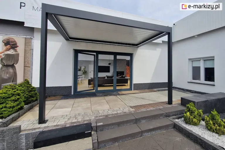 Nowoczesna pergola tarasowa przyścienna Sb400 w kolorze antracytowym, zmontowana na ekspozycji zewnętrznej. Konstrukcja posiada zadaszenie z białych lameli ustawionych w pozycji zamkniętej, pod którymi zamontowano liniowe oświetlenie LED biegnące wzdłuż wewnętrznych krawędzi ramy. Pergola osłania wejście do budynku z dużymi, przeszklonymi drzwiami tarasowymi w ciemnej ramie, przez które widać urządzone wnętrze salonu z szarą sofą i kominkiem. Podłoże pod pergolą jest zróżnicowane i składa się z dużych płyt tarasowych, kostki brukowej oraz ciemnych stopni schodowych. Po bokach widoczne są elementy dekoracyjne: niskie iglaki w pasie białego gresu oraz fragment baneru reklamowego z postacią kobiety. Zdjęcie prezentuje estetykę nocnego podświetlenia pergoli w zestawieniu z nowoczesną architekturą.