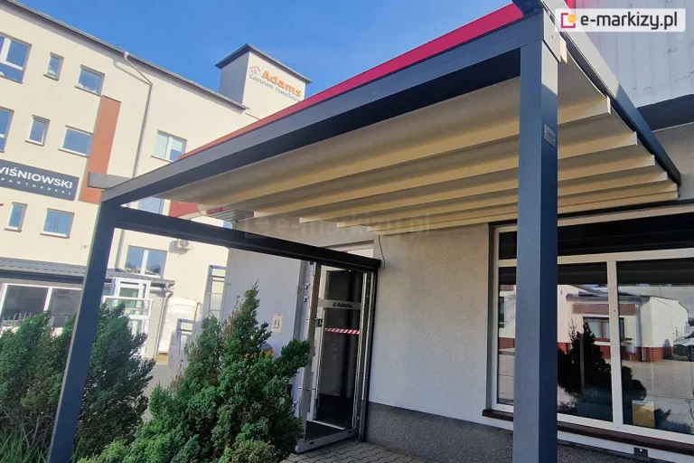 Zdjęcie przedstawia przyścienną pergolę tarasową Luxo z dachem rzymskim, zamontowaną nad wejściem do budynku komercyjnego. Konstrukcja nośna wykonana jest z profili aluminiowych w kolorze ciemnego antracytu i składa się z ramy zamocowanej do elewacji oraz przednich słupów o kwadratowym przekroju. Zadaszenie posiada ruchome poszycie z jasnej, kremowej tkaniny technicznej, która jest w pełni rozłożona i napięta na poprzecznych profilach, tworząc lekki skos odprowadzający wodę. Nad tkaniną, przy samej ścianie budynku, widoczny jest metalowy daszek ochronny w kolorze czerwonym, zabezpieczający mechanizm zwijający. Pergola znajduje się tuż obok wejścia z przeszklonymi drzwiami aluminiowymi i osłania duże okno z białymi ramami. Przed budynkiem znajduje się nawierzchnia z szarej kostki brukowej oraz niskie, zielone krzewy ozdobne. W tle widoczny jest wielopiętrowy budynek biurowy o jasnej elewacji z logo firm oraz bezchmurne, błękitne niebo.