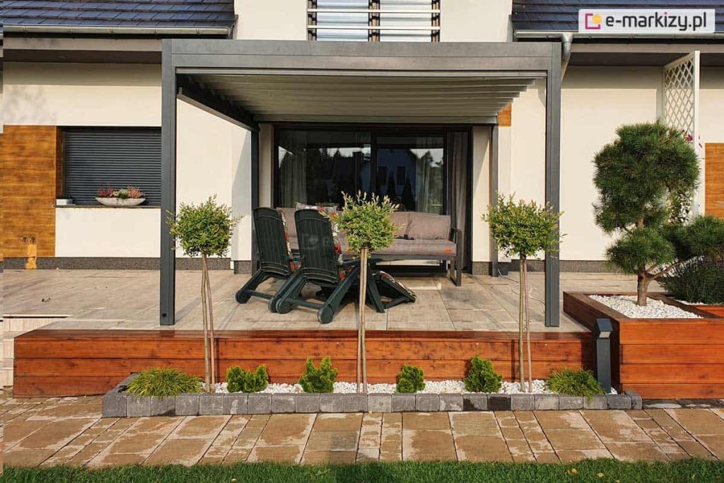 Zdjęcie prezentuje pergolę tarasową Luxury w kolorze antracytowym, zainstalowaną na podwyższonym tarasie wykonanym z jasnych płyt. Konstrukcja zadaszenia opiera się na dwóch solidnych słupach frontowych i jest zamontowana do białej elewacji domu, którą zdobią wstawki z paneli drewnopodobnych. Dach pergoli składa się z wodoodpornej tkaniny w białym kolorze, która jest rozwinięta na całym wysięgu, rzucając delikatny cień na strefę wypoczynkową. Pod pergolą znajduje się szara, nowoczesna sofa oraz dwa ciemnozielone fotele ogrodowe ustawione tyłem do ogrodu. Taras jest odseparowany od niższej części posesji drewnianym murkiem oporowym, przy którym posadzono ozdobne rośliny na pniu oraz niskie krzewy iglaste w białym żwirku. W tle widoczny jest fragment ogrodu z drzewami oraz sąsiednia zabudowa jednorodzinna. Całość kompozycji jest symetryczna i podkreśla nowoczesny charakter architektury budynku oraz jego otoczenia.