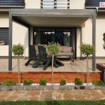 Zdjęcie prezentuje pergolę tarasową Luxury w kolorze antracytowym, zainstalowaną na podwyższonym tarasie wykonanym z jasnych płyt. Konstrukcja zadaszenia opiera się na dwóch solidnych słupach frontowych i jest zamontowana do białej elewacji domu, którą zdobią wstawki z paneli drewnopodobnych. Dach pergoli składa się z wodoodpornej tkaniny w białym kolorze, która jest rozwinięta na całym wysięgu, rzucając delikatny cień na strefę wypoczynkową. Pod pergolą znajduje się szara, nowoczesna sofa oraz dwa ciemnozielone fotele ogrodowe ustawione tyłem do ogrodu. Taras jest odseparowany od niższej części posesji drewnianym murkiem oporowym, przy którym posadzono ozdobne rośliny na pniu oraz niskie krzewy iglaste w białym żwirku. W tle widoczny jest fragment ogrodu z drzewami oraz sąsiednia zabudowa jednorodzinna. Całość kompozycji jest symetryczna i podkreśla nowoczesny charakter architektury budynku oraz jego otoczenia.