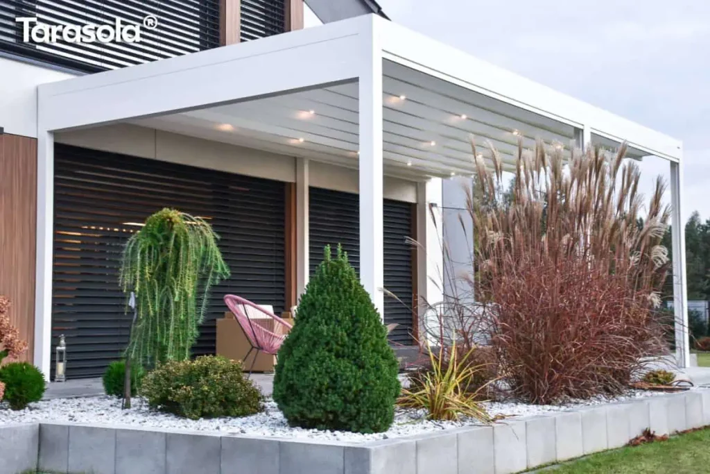 Wizualizacja przedstawia białą pergolę tarasową Tarasola Flat o prostej, nowoczesnej konstrukcji, zamontowaną bezpośrednio przy elewacji budynku. Zadaszenie wykonane jest z jasnej, rzymskiej tkaniny, która po rozłożeniu tworzy płaską powierzchnię z delikatnymi przetłoczeniami, w których zintegrowano punktowe oświetlenie LED o ciepłej barwie. Biała rama pergoli o matowym wykończeniu opiera się na smukłych słupach, tworząc zadaszoną przestrzeń nad tarasem. Pod konstrukcją znajduje się strefa wypoczynkowa z nowoczesnym, różowym fotelem typu "spaghetti". Taras oddzielony jest od ogrodu niskim murkiem z betonowych bloków, za którym wysypano biały żwir ozdobny i posadzono różnorodną roślinność, w tym wysokie trawy ozdobne o brązowych kłosach oraz formowane krzewy iglaste. W tle widoczna jest elewacja domu z ciemnymi, poziomymi żaluzjami fasadowymi, co nadaje całości elegancki i minimalistyczny charakter.