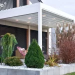 Wizualizacja przedstawia białą pergolę tarasową Tarasola Flat o prostej, nowoczesnej konstrukcji, zamontowaną bezpośrednio przy elewacji budynku. Zadaszenie wykonane jest z jasnej, rzymskiej tkaniny, która po rozłożeniu tworzy płaską powierzchnię z delikatnymi przetłoczeniami, w których zintegrowano punktowe oświetlenie LED o ciepłej barwie. Biała rama pergoli o matowym wykończeniu opiera się na smukłych słupach, tworząc zadaszoną przestrzeń nad tarasem. Pod konstrukcją znajduje się strefa wypoczynkowa z nowoczesnym, różowym fotelem typu "spaghetti". Taras oddzielony jest od ogrodu niskim murkiem z betonowych bloków, za którym wysypano biały żwir ozdobny i posadzono różnorodną roślinność, w tym wysokie trawy ozdobne o brązowych kłosach oraz formowane krzewy iglaste. W tle widoczna jest elewacja domu z ciemnymi, poziomymi żaluzjami fasadowymi, co nadaje całości elegancki i minimalistyczny charakter.