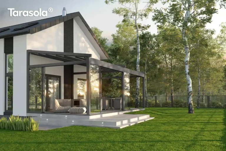 Nowoczesna pergola tarasowa Tarasola Elegancy w formie ogrodu zimowego, zamontowana do białej elewacji domu z dwuspadowym dachem. Konstrukcja pergoli w kolorze antracytowym posiada w pełni przeszklone ściany boczne w systemie przesuwnym oraz zadaszenie tkaninowe rzymskie, które jest rozłożone. Wewnątrz przeszklonej zabudowy znajduje się strefa relaksu z jasnymi, miękkimi pufami oraz stolikiem kawowym, a w dalszej części widoczny jest zestaw mebli jadalnianych. Taras został wykonany z jasnych płyt betonowych z wbudowanym punktowym oświetleniem LED w stopniach schodowych. Budynek posiada charakterystyczne ciemne pionowe pasy na elewacji, które komponują się z kolorem ramy pergoli. Całość zlokalizowana jest w otoczeniu dużego, zielonego trawnika oraz wysokich brzóz, za którymi widoczne jest metalowe ogrodzenie panelowe. Światło słoneczne odbija się w szklanych taflach zabudowy, podkreślając transparentność konstrukcji.