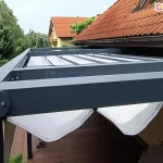Pergola rzymska Tako przymocowana do ściany budynku tuz pod spadzistym dachem pokrytym czerwoną dachówką. Aluminiowa konstrukcja pomalowana na kolor antracytowy kontrastuje z białą tkaniną. Pd pergolą widoczny fragment tarasu wyłożonego drewnianymi deskami, po prawej odwrócony do ściany mężczyzna, pod dachem widoczne dwie stopy w butach sportowych. Ponad pergolą widoczne liczne zielone drzewa oraz fragment sąsiedniego budynku.