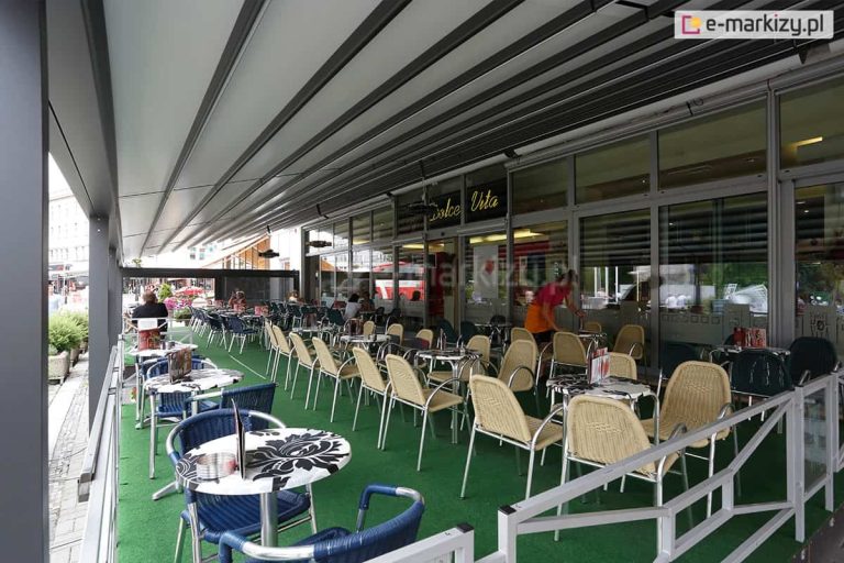 Zdjęcie prezentuje zastosowanie pergoli rzymskiej model Solid Selt jako zadaszenia ogródka restauracyjnego "Dolce Vita". Konstrukcja o imponującej rozpiętości wykonana jest z profili aluminiowych w kolorze antracytowym i została zamontowana wzdłuż przeszklonej fasady lokalu. Zadaszenie stanowi jasna, segmentowa tkanina, która jest całkowicie rozłożona, chroniąc dużą powierzchnię stolików przed słońcem lub deszczem. Przestrzeń pod pergolą jest wyposażona w liczne zestawy kawiarniane: stoliki z blatami w biało-czarne wzory kwiatowe oraz krzesła w kolorach beżowym i granatowym. Podłoże wyłożone jest estetyczną, zieloną wykładziną imitującą trawę. Ogródek jest oddzielony od chodnika niską, białą barierką o geometrycznym wzorze. Wewnątrz lokalu, za dużymi witrynami, widoczna jest obsługa oraz nowoczesne oświetlenie wnętrza. Perspektywa zdjęcia ukazuje rytmiczny układ profili dachu, który optycznie wydłuża przestrzeń restauracyjną.