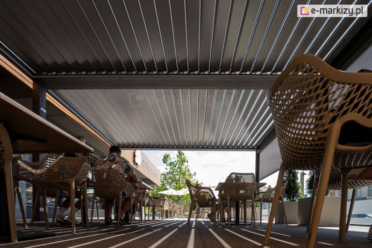 Widok z poziomu gruntu na wnętrze rozległej pergoli bioklimatycznej zainstalowanej w ogródku restauracyjnym. Zadaszenie składa się z gęsto rozmieszczonych, aluminiowych lameli w kolorze antracytowym, które są ustawione w pozycji lekko uchylonej, zapewniając ruch powietrza pod pergolą i osłonę przed mocnym słońcem. Pod pergolą znajdują się liczne stoliki oraz nowoczesne, ażurowe krzesła w kolorze beżowym o charakterystycznym splocie przypominającym siatkę. Na podłożu z kostki brukowej widoczne są wyraźne, równoległe pasy cienia i światła, powstające w przestrzeniach pomiędzy uchylonymi lamelami. W tle, pod zadaszeniem, siedzą goście restauracji, a w oddali widać słoneczny plac z drzewami liściastymi, białymi parasolami i fragmentami miejskiej zabudowy.