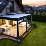 Zdjęcie wykonane z wysokiej perspektywy ukazuje nowoczesną zabudowę tarasową Essential tarasola w porze wieczornej, zamontowaną przy bocznej ścianie piętrowego domu jednorodzinnego. Konstrukcja o ciemnoantracytowym stelażu posiada płaski dach z białych lameli, który od spodu jest intensywnie doświetlony zintegrowanym systemem LED, rzucającym ciepły blask na całą przestrzeń użytkową. Taras, wyłożony jasną kostką brukową z dekoracyjnym obramowaniem, jest podzielony na dwie strefy: jadalnianą po lewej stronie z białym stołem i krzesłami na geometrycznym dywanie oraz wypoczynkową po prawej z jasną sofą i stolikiem kawowym. Pergola przylega do ściany z dużymi przeszkleniami, a powyżej widoczny jest balkon z ciemną balustradą oraz fragment dwuspadowego dachu pokrytego ciemną dachówką. Teren wokół tarasu obejmuje szeroki, zielony trawnik, ścieżkę z kostki oraz wysoki, gęsty żywopłot odgradzający posesję. W tle widoczna jest dalsza zabudowa osiedla oraz subtelne kolory zachodzącego słońca na horyzoncie, co kontrastuje z wyraźnym oświetleniem wnętrza pergoli.