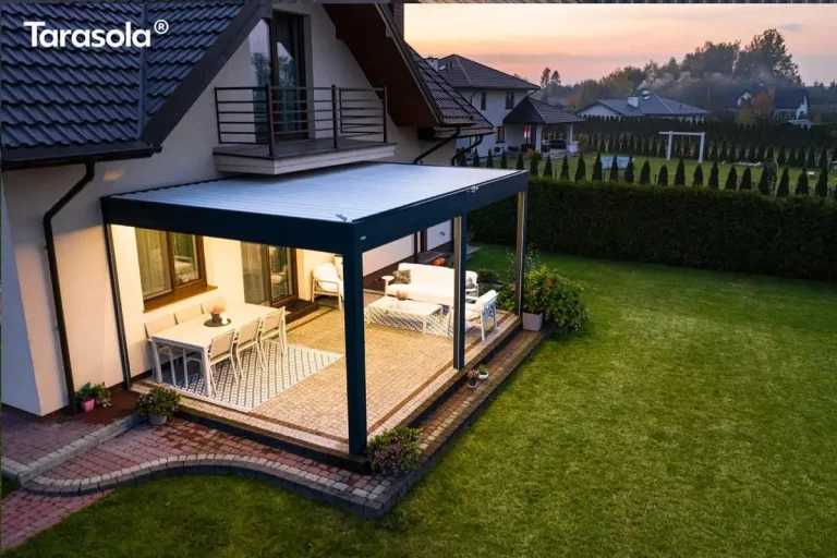 Zdjęcie wykonane z wysokiej perspektywy ukazuje nowoczesną zabudowę tarasową Essential tarasola w porze wieczornej, zamontowaną przy bocznej ścianie piętrowego domu jednorodzinnego. Konstrukcja o ciemnoantracytowym stelażu posiada płaski dach z białych lameli, który od spodu jest intensywnie doświetlony zintegrowanym systemem LED, rzucającym ciepły blask na całą przestrzeń użytkową. Taras, wyłożony jasną kostką brukową z dekoracyjnym obramowaniem, jest podzielony na dwie strefy: jadalnianą po lewej stronie z białym stołem i krzesłami na geometrycznym dywanie oraz wypoczynkową po prawej z jasną sofą i stolikiem kawowym. Pergola przylega do ściany z dużymi przeszkleniami, a powyżej widoczny jest balkon z ciemną balustradą oraz fragment dwuspadowego dachu pokrytego ciemną dachówką. Teren wokół tarasu obejmuje szeroki, zielony trawnik, ścieżkę z kostki oraz wysoki, gęsty żywopłot odgradzający posesję. W tle widoczna jest dalsza zabudowa osiedla oraz subtelne kolory zachodzącego słońca na horyzoncie, co kontrastuje z wyraźnym oświetleniem wnętrza pergoli.