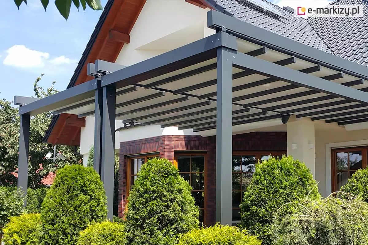 Zdjęcie prezentuje szeroką pergolę tarasową model Solid Aluprof o konstrukcji aluminiowej w kolorze ciemnego antracytu, zamontowaną do elewacji dużego domu jednorodzinnego. Zadaszenie składa się z dwóch niezależnych modułów tkaninowych w kolorze jasnokremowym, które są rozpięte na systemie poprzecznych profili aluminiowych. Konstrukcja wspiera się na trzech masywnych słupach frontowych o przekroju kwadratowym. Budynek posiada jasną elewację, fragmenty wykończone czerwoną cegłą klinkierową oraz ciemną dachówkę. Pergola osłania przestronny taras, a w jej bezpośrednim sąsiedztwie znajdują się gęste, formowane krzewy ozdobne (tuje) oraz inna niska roślinność ogrodowa. Całość kompozycji ukazana jest w pełnym świetle dziennym na tle bezchmurnego nieba, co podkreśla nowoczesny charakter instalacji i jej integrację z architekturą domu.