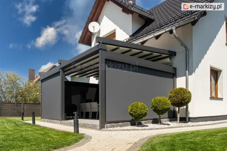 Zdjęcie prezentuje nowoczesną pergolę tarasową model Solid Selt w kolorze antracytowym, zainstalowaną przy białej elewacji domu jednorodzinnego o ciemnym, skośnym dachu. Kluczowym elementem tej realizacji są pionowe osłony boczne w postaci refleksoli Ziiip Selt w ciemnoszarym odcieniu, które są całkowicie opuszczone na dwóch sąsiadujących ścianach konstrukcji, zapewniając pełną prywatność i ochronę przed słońcem. Przez otwartą część frontową widoczny jest fragment wyposażenia tarasu, w tym plecione krzesła ogrodowe. Pergola posiada segmentowy, ruchomy dach z jasnej tkaniny, który w tej chwili jest częściowo zwinięty pod systemowy daszek ochronny. Taras wyłożony jest szarymi płytami i wyniesiony ponad poziom ogrodu, wykończony obrzeżem z łupanego kamienia. Wokół konstrukcji znajdują się dekoracyjne nasadzenia w postaci trzech krzewów formowanych w kule na niskich pniach, rosnących na pasie wysypanym jasnym żwirem. Całość otacza zadbany trawnik i chodnik z kostki brukowej pod bezchmurnym, błękitnym niebem.