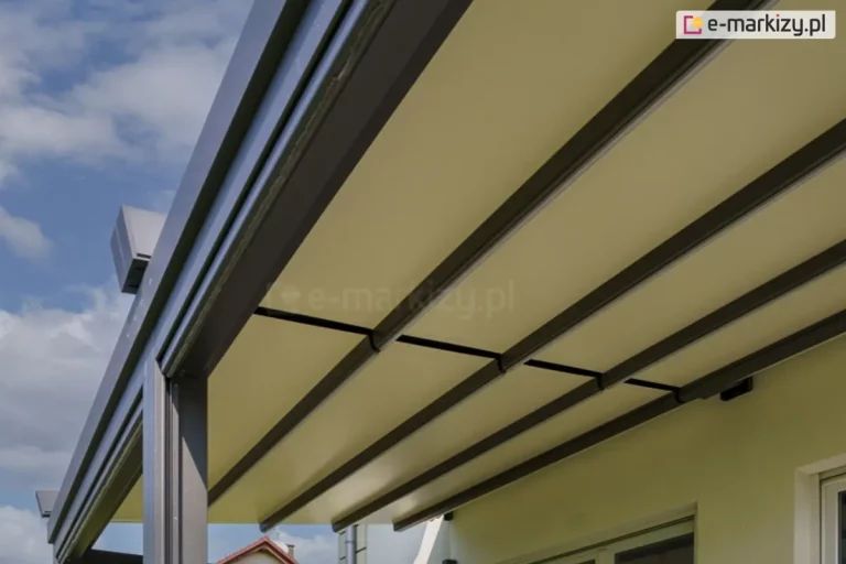 zbliżenie na dolną część zadaszenia pergoli model Solid Selt w widoku od strony tarasu. Konstrukcja zadaszenia opiera się na równolegle rozmieszczonych profilach poprzecznych w kolorze ciemnego antracytu, które podtrzymują gładko napiętą tkaninę w kolorze jasnokremowym. Materiał jest całkowicie rozłożony, tworząc jednolitą i płaską powierzchnię chroniącą przed słońcem. Po lewej stronie widoczny jest masywny profil boczny z prowadnicą, w której poruszają się elementy dachu. W górnym lewym rogu widać fragment zakończenia szyny jezdnej o geometrycznym kształcie. Tło stanowi jasna, tynkowana elewacja budynku z fragmentem ramy okiennej oraz błękitne niebo z rozproszonymi, białymi chmurami. Perspektywa zdjęcia z dołu podkreśla rytmiczny układ profili konstrukcyjnych i precyzyjne napięcie poszycia.