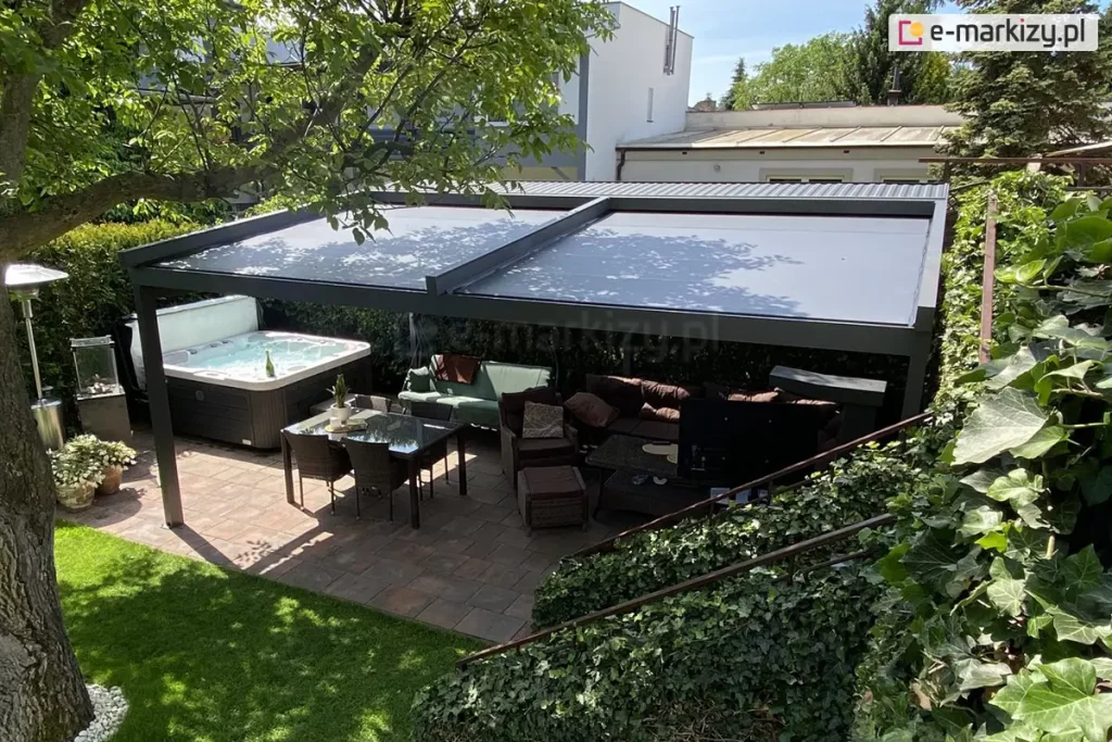 Zdjęcie prezentuje luksusową strefę wypoczynkową w ogrodzie, osłoniętą nowoczesną pergolą Square MOL o konstrukcji aluminiowej w kolorze antracytowym. Na górze pergoli zamontowano dwumodułowy dach tkaninowy z ciemnoszarym, gładkim poszyciem, które jest w pełni rozwinięte, zapewniając głęboki cień nad całym tarasem. Pod zadaszeniem znajduje się kompleksowo wyposażona przestrzeń rekreacyjna: po lewej stronie umieszczono duże, nowoczesne jacuzzi z białą niecką i ciemną obudową, natomiast centralną część zajmuje zestaw mebli ogrodowych, w tym narożna sofa z licznymi poduszkami oraz stół jadalniany z szklanym blatem i wiklinowymi krzesłami. Na tarasie widoczny jest również zewnętrzny grzejnik gazowy oraz donice z roślinami ozdobnymi. Nawierzchnia tarasu została wykonana z wielkoformatowych płyt kamiennych w odcieniach brązu i szarości. Pergola jest otoczona gęstą roślinnością, w tym wysokim żywopłotem oraz dużym liściastym drzewem po lewej stronie, którego gałęzie częściowo nachodzą nad konstrukcję. W tle widoczne są dachy sąsiednich budynków oraz jasne, bezchmurne niebo, co podkreśla letni, wypoczynkowy charakter aranżacji.