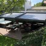 Zdjęcie prezentuje luksusową strefę wypoczynkową w ogrodzie, osłoniętą nowoczesną pergolą Square MOL o konstrukcji aluminiowej w kolorze antracytowym. Na górze pergoli zamontowano dwumodułowy dach tkaninowy z ciemnoszarym, gładkim poszyciem, które jest w pełni rozwinięte, zapewniając głęboki cień nad całym tarasem. Pod zadaszeniem znajduje się kompleksowo wyposażona przestrzeń rekreacyjna: po lewej stronie umieszczono duże, nowoczesne jacuzzi z białą niecką i ciemną obudową, natomiast centralną część zajmuje zestaw mebli ogrodowych, w tym narożna sofa z licznymi poduszkami oraz stół jadalniany z szklanym blatem i wiklinowymi krzesłami. Na tarasie widoczny jest również zewnętrzny grzejnik gazowy oraz donice z roślinami ozdobnymi. Nawierzchnia tarasu została wykonana z wielkoformatowych płyt kamiennych w odcieniach brązu i szarości. Pergola jest otoczona gęstą roślinnością, w tym wysokim żywopłotem oraz dużym liściastym drzewem po lewej stronie, którego gałęzie częściowo nachodzą nad konstrukcję. W tle widoczne są dachy sąsiednich budynków oraz jasne, bezchmurne niebo, co podkreśla letni, wypoczynkowy charakter aranżacji.