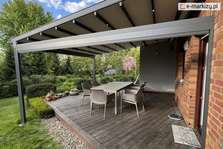 Nowoczesna pergola tarasowa Square o konstrukcji aluminiowej w kolorze ciemnoszarym, zamontowana do elewacji budynku wykonanej z czerwonej cegły oraz szarego tynku. Zadaszenie pergoli wyposażone jest w zintegrowane oświetlenie punktowe LED, umieszczone w poprzecznych profilach podtrzymujących jasne poszycie materiałowe. Konstrukcja wsparta jest na solidnych, pionowych słupach o kwadratowym przekroju zamocowanych w trawniku, a jej przednia belka posiada system odprowadzania wody. Pod pergolą, na tarasie z ciemnych desek drewnianych układanych pod skosem, znajduje się zestaw mebli składający się z dużego, szarego stołu oraz sześciu dopasowanych kolorystycznie krzeseł z technorattanu. W rogu tarasu ustawiono rząd donic z drobnymi kwiatami w kolorach żółtym i białym. Przy wejściu do domu na deski rzucono parę ciemnych klapek oraz wycieraczkę z geometrycznym wzorem. Tło stanowi bogata roślinność ogrodowa z przystrzyżonymi krzewami, wysokimi drzewami iglastymi oraz kwitnącym na różowo drzewem. Całość ujęta jest w świetle dziennym pod błękitnym niebem z rozproszonymi chmurami.