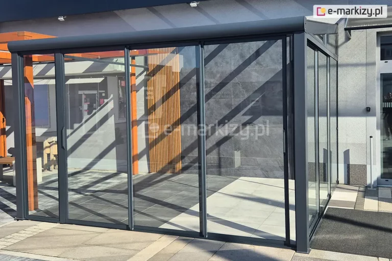 Nowoczesna pergola szklana o konstrukcji aluminiowej w kolorze antracytowym, zamontowana przy elewacji budynku. Obiekt posiada zintegrowany system przesuwnych drzwi szklanych, które tworzą szczelną zabudowę ścian bocznych i frontowej. Panele szklane są osadzone w cienkich, ciemnych profilach, a na skrajnych skrzydłach widoczne są pionowe uchwyty ułatwiające przesuwanie tafli. Dach konstrukcji jest nachylony i wykonany z przezroczystego szkła, wsparty na solidnych słupach nośnych o kwadratowym przekroju. Wewnątrz zabudowy widoczna jest posadzka wykonana z wielkoformatowych płyt tarasowych w dwóch odcieniach szarości. Tło stanowi fragment elewacji budynku wyłożony ciemnoszarymi płytami o strukturze kamienia oraz pionowe lamele drewniane w naturalnym kolorze. Na powierzchni szyb i podłożu widoczny jest wyraźny rysunek cieni rzucanych przez konstrukcję w słoneczny dzień. Obok pergoli znajduje się fragment innej konstrukcji drewnianej oraz przeszklone wejście do budynku. Całość zabudowy jest posadowiona na płaskim, utwardzonym podłożu z kostki brukowej i płyt chodnikowych.