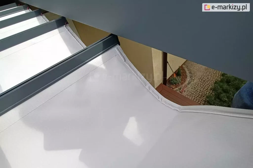 Widok od góry na tkaninę dachu Tako, z widocznym zaszyciem przewodów do oświetlenia LED w krawędzi białej tkaniny mocowanej do antracytowej belki przeciwwietrznej. W przerwie pomiędzy tkaniną a konstrukcją widoczny fragment tarasu z drewnianym podestem oraz fragment wyłożony kostką brukową, na którym stoi oparta o ścianę miotła.