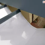 Widok od góry na tkaninę dachu Tako, z widocznym zaszyciem przewodów do oświetlenia LED w krawędzi białej tkaniny mocowanej do antracytowej belki przeciwwietrznej. W przerwie pomiędzy tkaniną a konstrukcją widoczny fragment tarasu z drewnianym podestem oraz fragment wyłożony kostką brukową, na którym stoi oparta o ścianę miotła.