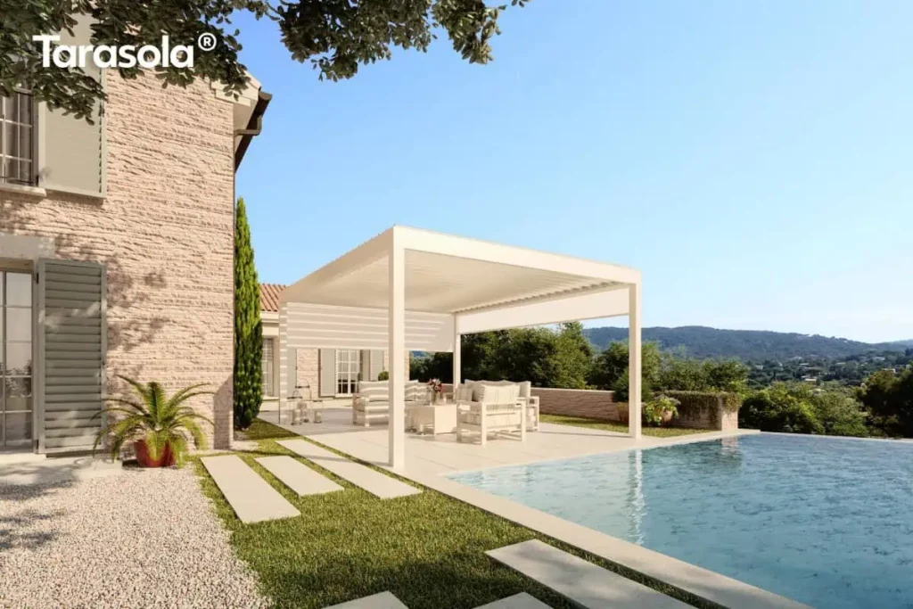 Nowoczesna pergola bioklimatyczna essential Tarasola w kolorze kremowym lub białym, zamontowana jako konstrukcja wolnostojąca na jasnym tarasie przy basenie typu infinity. Produkt charakteryzuje się płaskim dachem z ruchomymi lamelami oraz zintegrowaną boczną osłoną przeciwsłoneczną typu screen w kolorze dopasowanym do stelaża, która jest częściowo rozwinięta, zapewniając prywatność i ochronę przed słońcem. Pod zadaszeniem znajduje się komplet białych mebli ogrodowych z miękkimi poduszkami, ustawiony na gładkich płytach tarasowych. Obok konstrukcji widoczna jest ściana budynku z jasnej cegły lub kamienia z klasycznymi, szarymi okiennicami oraz starannie zaplanowana zieleń w formie trawnika, ścieżki z płyt kamiennych i roślin doniczkowych. W tle rozpościera się szeroka panorama na zalesione wzgórza pod czystym, błękitnym niebem, co podkreśla luksusowy charakter instalacji w otoczeniu natury.