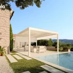 Nowoczesna pergola bioklimatyczna essential Tarasola w kolorze kremowym lub białym, zamontowana jako konstrukcja wolnostojąca na jasnym tarasie przy basenie typu infinity. Produkt charakteryzuje się płaskim dachem z ruchomymi lamelami oraz zintegrowaną boczną osłoną przeciwsłoneczną typu screen w kolorze dopasowanym do stelaża, która jest częściowo rozwinięta, zapewniając prywatność i ochronę przed słońcem. Pod zadaszeniem znajduje się komplet białych mebli ogrodowych z miękkimi poduszkami, ustawiony na gładkich płytach tarasowych. Obok konstrukcji widoczna jest ściana budynku z jasnej cegły lub kamienia z klasycznymi, szarymi okiennicami oraz starannie zaplanowana zieleń w formie trawnika, ścieżki z płyt kamiennych i roślin doniczkowych. W tle rozpościera się szeroka panorama na zalesione wzgórza pod czystym, błękitnym niebem, co podkreśla luksusowy charakter instalacji w otoczeniu natury.