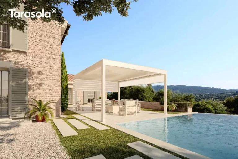 Nowoczesna pergola bioklimatyczna essential Tarasola w kolorze kremowym lub białym, zamontowana jako konstrukcja wolnostojąca na jasnym tarasie przy basenie typu infinity. Produkt charakteryzuje się płaskim dachem z ruchomymi lamelami oraz zintegrowaną boczną osłoną przeciwsłoneczną typu screen w kolorze dopasowanym do stelaża, która jest częściowo rozwinięta, zapewniając prywatność i ochronę przed słońcem. Pod zadaszeniem znajduje się komplet białych mebli ogrodowych z miękkimi poduszkami, ustawiony na gładkich płytach tarasowych. Obok konstrukcji widoczna jest ściana budynku z jasnej cegły lub kamienia z klasycznymi, szarymi okiennicami oraz starannie zaplanowana zieleń w formie trawnika, ścieżki z płyt kamiennych i roślin doniczkowych. W tle rozpościera się szeroka panorama na zalesione wzgórza pod czystym, błękitnym niebem, co podkreśla luksusowy charakter instalacji w otoczeniu natury.