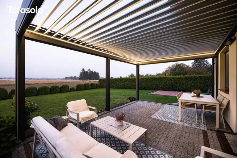 Zdjęcie prezentuje perspektywę z narożnika tarasu, ukazującą pełną funkcjonalność i nowoczesny design konstrukcji Essential tarasola o zmierzchu. Na pierwszym planie znajduje się biała sofa ogrodowa z ciemną poduszką oraz biały stolik kawowy, ustawione na niebiesko-białym dywanie o geometrycznym wzorze. Po prawej stronie, na drugim dywanie, widoczny jest biały stół jadalniany z kompletem krzesł. Konstrukcja zadaszenia w kolorze antracytowym posiada dach z białych lameli, które są częściowo uchylone, a ich krawędzie są efektownie podświetlone zintegrowanym systemem oświetlenia LED o ciepłej barwie. Pergola jest otwarta z dwóch stron, co zapewnia swobodny widok na przestronny, zielony ogród, niski żywopłot z kulistych krzewów oraz rozległe pola pod jasnym niebem. Podłoga tarasu wyłożona jest drobną, jasnoszarą kostką brukową z ciemniejszymi pasami dekoracyjnymi. Całość kompozycji oddaje atmosferę spokoju i luksusu, podkreślając harmonijne połączenie wnętrza domu z przestrzenią ogrodu.