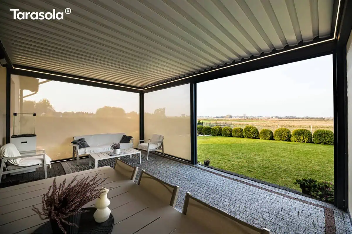 Zdjęcie prezentuje perspektywę z wnętrza luksusowego tarasu, osłoniętego nowoczesną pergolą Essential tarasola. Na pierwszym planie widoczny jest fragment jasnego, prostokątnego stołu jadalnianego, na którym ustawiono dekoracyjną roślinę w ciemnej donicy oraz białą świecę o nieregularnym kształcie. Konstrukcja zadaszenia o barwie głębokiego antracytu posiada dach z białych, aluminiowych lameli, które są w pełni zamknięte, oraz zintegrowane liniowe oświetlenie LED biegnące wzdłuż wewnętrznych krawędzi ramy. Po lewej stronie oraz w części środkowej zamontowano rolety typu screen w kolorze beżowym, które są całkowicie opuszczone, tworząc intymną przestrzeń wypoczynkową z białą sofą, stolikiem kawowym i fotelem. Prawa strona pergoli pozostaje otwarta, ukazując rozległy, intensywnie zielony trawnik, rządek niskich, formowanych w kule krzewów oraz szeroką panoramę pól pod jasnym niebem. Podłoga tarasu została wykonana z szarej kostki brukowej, a cała aranżacja łączy w sobie cechy minimalistycznej architektury z przytulnym, ogrodowym klimatem.