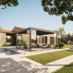 Nowoczesna pergola wolnostojąca essential Tarasola w kolorze ciemnoszarym antacytowym, zainstalowana na przestronnym tarasie z jasnych płyt kamiennych obok basenu. Konstrukcja posiada płaski dach z ruchomymi lamelami oraz szklane zabudowy boczne w formie przesuwnych paneli, które tworzą zamkniętą, transparentną bryłę ogrodową. Wewnątrz pergoli znajduje się strefa wypoczynkowa z jasnoszarą sofą, fotelami oraz niskim stolikiem kawowym ustawionym na dekoracyjnym żwirze. Pergola przylega wizualnie do parterowego budynku o elewacji wykonanej z naturalnego, jasnego kamienia z brązowymi okiennicami. W tle widoczny jest zadbany trawnik, kamienny mur porośnięty roślinnością oraz liczne drzewa liściaste pod bezchmurnym, błękitnym niebem. Scena przedstawia luksusową aranżację przestrzeni zewnętrznej, w której aluminiowa konstrukcja harmonizuje z klasyczną architekturą domu i elementami małej architektury ogrodowej.