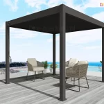 Pergola aluminiowa z dachem lamelowym Marlo w kolorze antracytowym, z napędem ręcznym korbowym, ustawiona na tarasie z jasnoszarych desek z widokiem na morze, skały i drzewa. Pod pergolą w pobliżu niskiej, szklanej barierki tarasu ustawiono trzy krzesła z plecionym oparciem, poduszkami na cienkich metalowych podporach.