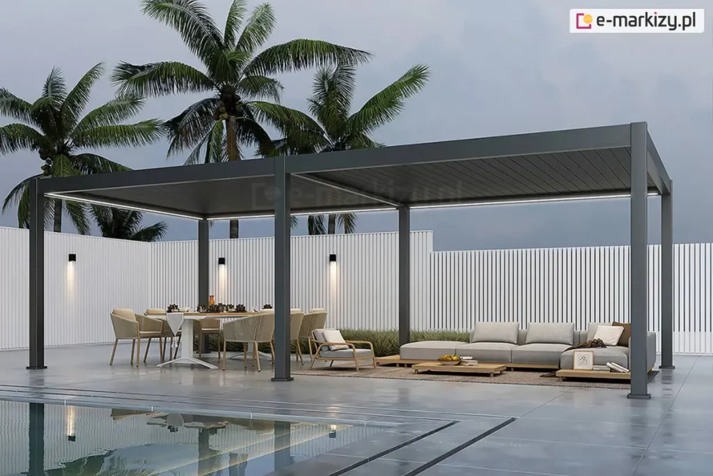 Wielkogabarytowa, dwumodułowa pergola bioklimatyczna Rubio o długości 8 metrów, wykonana z aluminium w kolorze antracytowym. Konstrukcja wsparta na sześciu masywnych słupach nośnych, tworzy rozległą, zadaszoną strefę rekreacyjną nad brzegiem basenu. Dach z lamelami w pozycji zamkniętej posiada zintegrowane oświetlenie liniowe LED o barwie chłodnej, poprowadzone wzdłuż wewnętrznych belek obu modułów. Pod pergolą wydzielono dwie strefy: jadalnianą z dużym stołem i sześcioma fotelami oraz wypoczynkową z niską, modułową sofą i stolikami kawowymi na dywanie tarasowym. Przestrzeń jest ograniczona wysokim ogrodzeniem z białych pionowych lameli, na którym zamontowano nowoczesne kinkiety świecące góra-dół. W tle widoczne są wysokie palmy kokosowe na tle zachmurzonego, stalowego nieba. Pierwszy plan zajmuje tafla lustrzanego basenu, w której odbija się konstrukcja pergoli oraz oświetlenie. Całość aranżacji utrzymana jest w prestiżowym stylu nowoczesnego resortu.