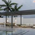 Wielkogabarytowa, dwumodułowa pergola bioklimatyczna Rubio o długości 8 metrów, wykonana z aluminium w kolorze antracytowym. Konstrukcja wsparta na sześciu masywnych słupach nośnych, tworzy rozległą, zadaszoną strefę rekreacyjną nad brzegiem basenu. Dach z lamelami w pozycji zamkniętej posiada zintegrowane oświetlenie liniowe LED o barwie chłodnej, poprowadzone wzdłuż wewnętrznych belek obu modułów. Pod pergolą wydzielono dwie strefy: jadalnianą z dużym stołem i sześcioma fotelami oraz wypoczynkową z niską, modułową sofą i stolikami kawowymi na dywanie tarasowym. Przestrzeń jest ograniczona wysokim ogrodzeniem z białych pionowych lameli, na którym zamontowano nowoczesne kinkiety świecące góra-dół. W tle widoczne są wysokie palmy kokosowe na tle zachmurzonego, stalowego nieba. Pierwszy plan zajmuje tafla lustrzanego basenu, w której odbija się konstrukcja pergoli oraz oświetlenie. Całość aranżacji utrzymana jest w prestiżowym stylu nowoczesnego resortu.
