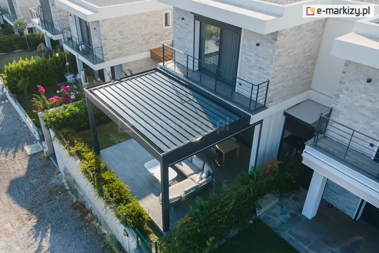 Widok z lotu ptaka na nowoczesną pergolę bioklimatyczną Sb400 o konstrukcji aluminiowej w kolorze antracytowym, zainstalowaną na tarasie luksusowego domu jednorodzinnego. Dach pergoli z zamkniętymi ciemnymi lamelami przylega do elewacji budynku wykonanej z jasnego kamienia dekoracyjnego i białego tynku. Pod zadaszeniem widoczny jest komplet jasnych mebli wypoczynkowych w kształcie litery L, biały stolik kawowy oraz drewniany stół jadalniany. Taras przylega do zadbanego ogrodu z zielonym żywopłotem i kolorowymi kwiatami. Na piętrze budynku znajduje się balkon z metalową, czarną barierką oraz przesuwne drzwi tarasowe. W tle widoczne są sąsiednie, bliźniacze budynki o podobnej architekturze, a po lewej stronie fragment drogi dojazdowej wysypanej żwirem.