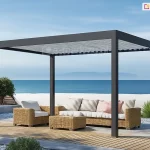 Wolnostojąca pergola bioklimatyczna Rubio w kolorze antracytowym, zlokalizowana na luksusowym tarasie nadmorskim. Dach wykonany z aluminiowych lameli w kolorze jasnoszarym jest ustawiony w pozycji otwartej, co pozwala na naturalną wentylację. Pod konstrukcją znajduje się strefa wypoczynkowa z meblami z naturalnego technorattanu: dużą sofą z jasnymi poduszkami dekoracyjnymi, fotelami oraz pasującym stolikiem kawowym. Nawierzchnia tarasu to połączenie klasycznej deski kompozytowej w odcieniu naturalnego drewna oraz jasnych płyt kamiennych otoczonych białym grysem dekoracyjnym. W bezpośrednim sąsiedztwie pergoli widoczna jest niska, biała ściana oddzielająca taras od plaży, za którą rozciąga się bezkresny widok na błękitne morze i zamglony horyzont pod czystym niebem. Roślinność ograniczona do minimalistycznych form: niskich krzewów oraz ozdobnych traw posadzonych w gruncie. Całość aranżacji podkreśla harmonijne połączenie nowoczesnej technologii osłonowej z naturalnym, nadmorskim otoczeniem.