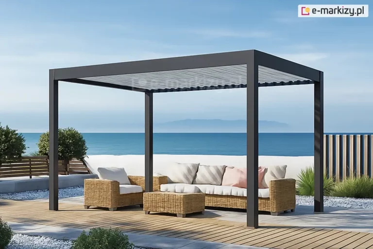 Wolnostojąca pergola bioklimatyczna Rubio w kolorze antracytowym, zlokalizowana na luksusowym tarasie nadmorskim. Dach wykonany z aluminiowych lameli w kolorze jasnoszarym jest ustawiony w pozycji otwartej, co pozwala na naturalną wentylację. Pod konstrukcją znajduje się strefa wypoczynkowa z meblami z naturalnego technorattanu: dużą sofą z jasnymi poduszkami dekoracyjnymi, fotelami oraz pasującym stolikiem kawowym. Nawierzchnia tarasu to połączenie klasycznej deski kompozytowej w odcieniu naturalnego drewna oraz jasnych płyt kamiennych otoczonych białym grysem dekoracyjnym. W bezpośrednim sąsiedztwie pergoli widoczna jest niska, biała ściana oddzielająca taras od plaży, za którą rozciąga się bezkresny widok na błękitne morze i zamglony horyzont pod czystym niebem. Roślinność ograniczona do minimalistycznych form: niskich krzewów oraz ozdobnych traw posadzonych w gruncie. Całość aranżacji podkreśla harmonijne połączenie nowoczesnej technologii osłonowej z naturalnym, nadmorskim otoczeniem.