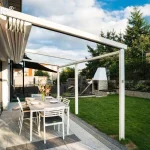 Biała pergola tarasowa Tarasola Simple o konstrukcji aluminiowej z całkowicie otwartym dachem, zamontowana do ściany budynku wykończonej panelami o strukturze drewna. Tkanina zadaszenia jest zwinięta w estetyczne fałdy przy elewacji, odsłaniając widok na niebo i aluminiowe szyny prowadzące. Konstrukcja opiera się na dwóch białych słupach nośnych, pod którymi na nowoczesnym tarasie z płyt ustawiono prostokątny stół jadalniany i sześć białych krzeseł. W tle widoczny jest zielony trawnik, murowany grill ogrodowy, kamienne ogrodzenie oraz wysokie drzewa iglaste pod lekko zachmurzonym niebem.