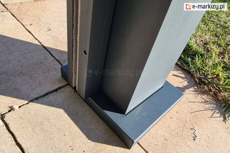 Zbliżenie na podstawę pionowego słupa nośnego nowoczesnej pergoli bioklimatycznej, zamontowanego na tarasie z jasnej kostki brukowej. Słup o kwadratowym przekroju oraz jego systemowa maskownica stopy montażowej wykonane są z aluminium malowanego proszkowo na kolor ciemnego antracytu o matowej, drobnoziarnistej strukturze. Maskownica o prostokątnym kształcie ściśle przylega do podłoża, estetycznie zakrywając elementy kotwiące konstrukcję do fundamentu. Do bocznej ścianki słupa przylega pionowy profil prowadnicy rolety zewnętrznej, wewnątrz którego widoczna jest szczotka uszczelniająca oraz pionowy kanał techniczny. W profilu prowadnicy znajduje się niewielki, fabrycznie wycięty otwór montażowy. Zdjęcie zostało wykonane w pełnym świetle dziennym, co uwydatnia detale wykończenia metalu oraz teksturę betonowej kostki brukowej, na którą pada wyraźny cień rzucany przez konstrukcję. W prawym górnym rogu kadru widoczny jest fragment zielonego trawnika.