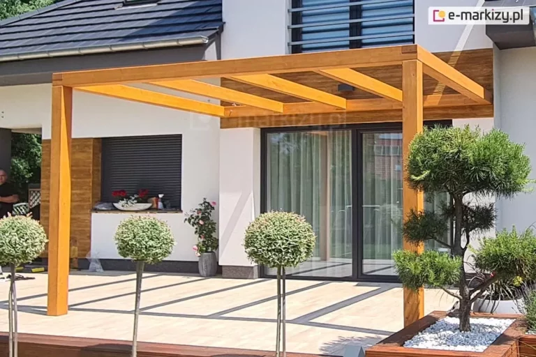 Pergola drewniana Juko przyścienna pokryta lakierobejcą ustawiona na tarasie z jasną płytką otoczonym roślinnością.