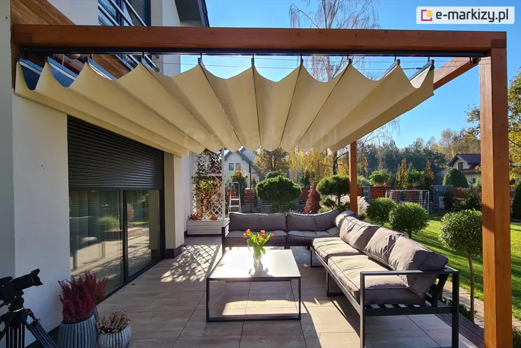 Pergola tarasowa drewniana koro z dachem rzymskim supro nad nasłonecznionym tarasem z meblami i widocznym w tle ogrodem.
