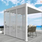 Wolnostojąca pergola tarasowa Marlo w kolorze białym, posadowiona na tarasie wyłożonym deską kompozytową w odcieniu jasnego drewna. Aluminiowa konstrukcja o prostokątnym przekroju profili charakteryzuje się zabudowaną ścianą boczną, którą tworzą trzy pionowe panele z poziomymi lamelami typu shutters, umożliwiającymi regulację dopływu światła i powietrza. Na lewym skrajnym profilu widoczny jest mechanizm korbowy służący do obsługi ruchomych elementów. Zadaszenie pergoli stanowią równolegle ułożone lamele dachowe w tym samym białym kolorze. Wewnątrz konstrukcji ustawione są dwa nowoczesne fotele z technorattanu z jasnymi poduszkami oraz niewielki stolik. Tło stanowi nadmorski krajobraz z błękitnym morzem, linią brzegową oraz bezchmurnym niebem, co podkreśla wypoczynkowy charakter produktu.