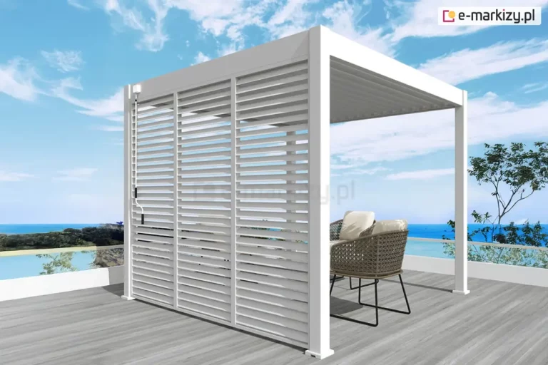 Wolnostojąca pergola tarasowa Marlo w kolorze białym, posadowiona na tarasie wyłożonym deską kompozytową w odcieniu jasnego drewna. Aluminiowa konstrukcja o prostokątnym przekroju profili charakteryzuje się zabudowaną ścianą boczną, którą tworzą trzy pionowe panele z poziomymi lamelami typu shutters, umożliwiającymi regulację dopływu światła i powietrza. Na lewym skrajnym profilu widoczny jest mechanizm korbowy służący do obsługi ruchomych elementów. Zadaszenie pergoli stanowią równolegle ułożone lamele dachowe w tym samym białym kolorze. Wewnątrz konstrukcji ustawione są dwa nowoczesne fotele z technorattanu z jasnymi poduszkami oraz niewielki stolik. Tło stanowi nadmorski krajobraz z błękitnym morzem, linią brzegową oraz bezchmurnym niebem, co podkreśla wypoczynkowy charakter produktu.