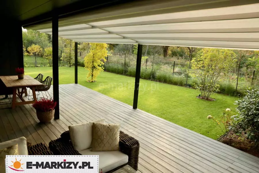 Widok z wnętrza tarasu na ogród, ukazujący spód zadaszenia pergola Light tarasola w pełnym rozłożeniu. Jasna, kremowa tkanina techniczna dachu jest napięta na poprzecznych profilach, tworząc płaską, lecz lekko pofalowaną powierzchnię, która skutecznie zacienia całą strefę wypoczynkową. Konstrukcja wsparta jest na smukłych, czarnych profilach aluminiowych, które harmonizują z nowoczesnym designem budynku. Pod zadaszeniem znajduje się komfortowa strefa relaksu wyposażona w meble technorattanowe z jasnymi poduszkami oraz drewniany stół jadalniany, na którym ustawiono donicę z czerwonymi kwiatami. Podłoga tarasu wykonana jest z szarych desek kompozytowych o wyraźnym usłojeniu. Perspektywa zdjęcia otwiera się na rozległy, intensywnie zielony trawnik, otoczony gęstymi nasadzeniami ozdobnych traw, krzewów i drzew liściastych. W oddali widoczne jest ażurowe ogrodzenie posesji, a cała scena skąpana jest w miękkim, rozproszonym świetle dziennym, co podkreśla przytulną i luksusową atmosferę wypoczynku pod pergolą Light tarasola.