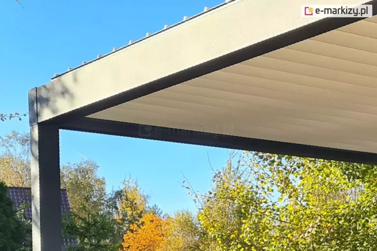 Zdjęcie prezentuje perspektywę od dołu na krawędź nowoczesnej pergoli bioklimatycznej, skupiając się na styku aluminiowej ramy z systemem lamelowego zadaszenia. Masywna belka konstrukcyjna o prostokątnym przekroju, wykończona matowym lakierem w kolorze ciemnego antracytu, wyznacza górną ramę budowli. Pod jej dolną krawędzią zamontowana jest estetyczna, liniowa listwa oświetleniowa LED, chroniona mlecznym kloszem, która biegnie wzdłuż profilu. Zadaszenie tworzą szerokie, aluminiowe lamele w kolorze białym, które na zdjęciu są całkowicie domknięte, tworząc gładką i szczelną płaszczyznę dachu. Widoczne są precyzyjnie dopasowane zakończenia lameli wykonane z białego tworzywa, które stykają się z czarną szyną prowadzącą ukrytą wewnątrz ramy. W tle, ponad linią dachu, rozpościera się bezchmurne, intensywnie niebieskie niebo, a poniżej ramy widoczne są gęste korony drzew o zielonych i lekko żółknących liściach. Ujęcie podkreśla nowoczesny design, czystość formy oraz wysoki standard technicznego wykończenia konstrukcji.