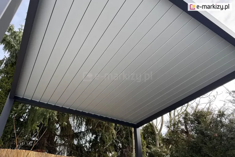 Szczelnie zamknięte lamele o jasnoszarej barwie osadzone na antracytowej konstrukcji pergoli Marlo tworzą skuteczne zadaszenie przed deszczem i słońcem. W tle widoczne niskie drzewa i krzewy iglaste.